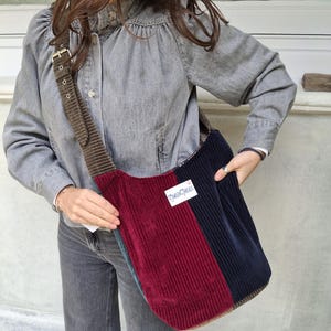 May include: A corduroy shoulder bag with a patchwork design. The bag features vertical panels in burgundy, navy, and teal, with a brown corduroy strap. A white label with text is visible on the front.