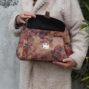 May include: A handbag featuring a floral print with a brown base and red and pink roses. It has a silver clasp and a black interior. The person holding the bag is wearing a cream-coloured coat.