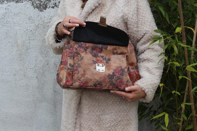 May include: A handbag featuring a floral print with a brown base and red and pink roses. It has a silver clasp and a black interior. The person holding the bag is wearing a cream-coloured coat.