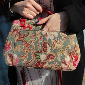 May include: A floral tapestry handbag with red leather accents and a red strap. The bag has a green background with a floral pattern in shades of pink, red, yellow, and brown.