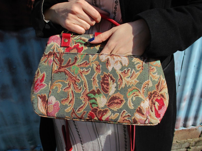 May include: A floral tapestry handbag with red leather accents and a red strap. The bag has a green background with a floral pattern in shades of pink, red, yellow, and brown.