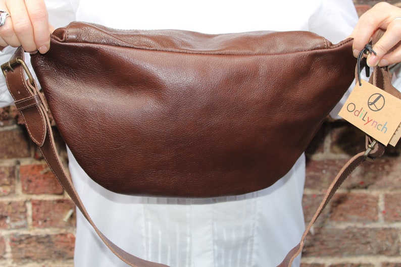 K&ouml;nnte beinhalten: Eine braune Leder-Bauchtasche mit einem langen verstellbaren Riemen. Die Tasche hat ein Etikett mit der Aufschrift "odiLynch" und einem stilisierten Vogel-Logo.