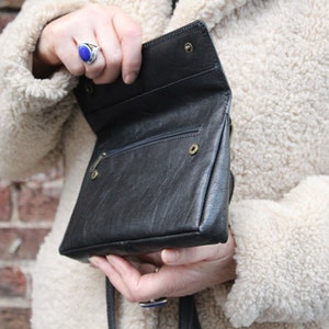 K&ouml;nnte beinhalten: Eine schwarze Leder-Umh&auml;ngetasche mit Klappenverschluss und Rei&szlig;verschlussfach. Die Tasche hat einen Riemen und ist ge&ouml;ffnet, um das Innere zu zeigen.