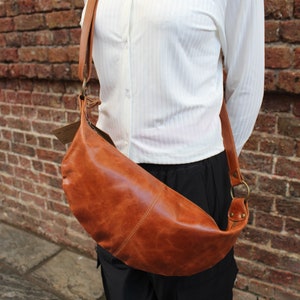 May include: A brown leather crossbody bag with a long strap. The bag is worn over the shoulder of a person wearing a white button-down shirt and black pants.