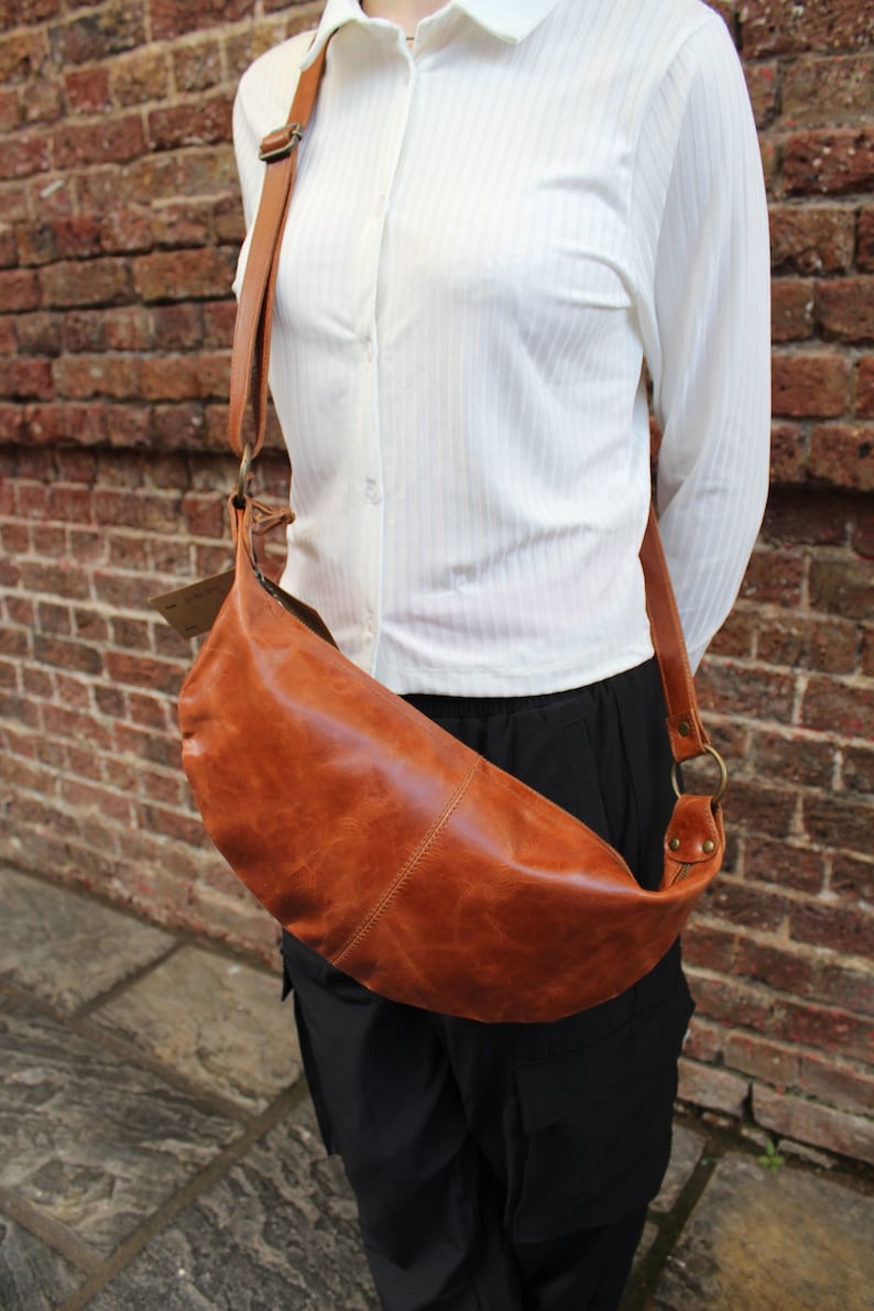 May include: A brown leather crossbody bag with a long strap. The bag is worn over the shoulder of a person wearing a white button-down shirt and black pants.