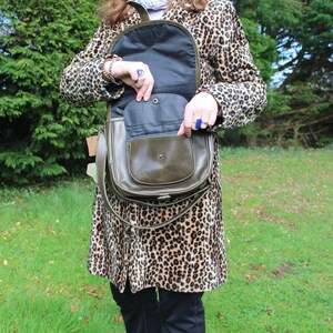 May include: A brown leather satchel bag with a flap closure and a long strap. The bag is open and the inside is lined with a dark fabric. The bag is being held by a person wearing a leopard print coat.