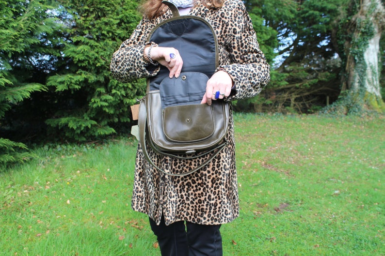 May include: A brown leather satchel bag with a flap closure and a long strap. The bag is open and the inside is lined with a dark fabric. The bag is being held by a person wearing a leopard print coat.