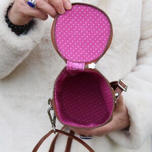 May include: Open, round brown purse with a zipper, revealing a pink interior with white polka dots. The purse has a brown strap and a silver clasp. The purse is held open, showcasing the interior design.