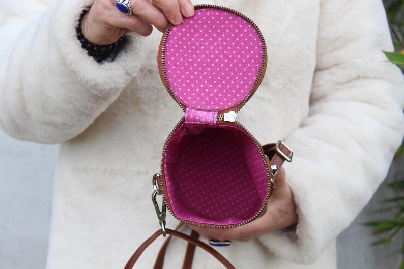 May include: Open, round brown purse with a zipper, revealing a pink interior with white polka dots. The purse has a brown strap and a silver clasp. The purse is held open, showcasing the interior design.