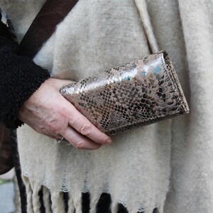 Könnte beinhalten: Eine Clutch mit Schlangenmuster in Braun- und Silbertönen wird vor einem cremefarbenen Schal mit Fransen gehalten. Die Clutch hat einen metallischen Glanz und eine rechteckige Form. Ein brauner Lederriemen ist sichtbar.