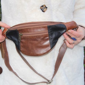 Könnte beinhalten: Eine braune Leder-Bauchtasche mit einem schwarzen Lederpanel und einer Reißverschlusstasche. Die Bauchtasche wird um die Taille getragen und hat einen langen Riemen.