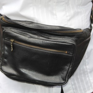 May include: Black leather fanny pack with multiple zippered compartments and gold-colored zippers. The bag is worn around the waist and has a rectangular shape. The fanny pack is a practical accessory for carrying essentials.