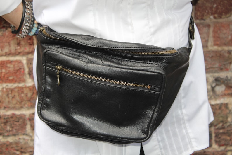 May include: Black leather fanny pack with multiple zippered compartments and gold-colored zippers. The bag is worn around the waist and has a rectangular shape. The fanny pack is a practical accessory for carrying essentials.