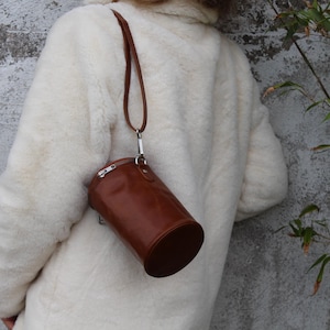 May include: A brown leather cylindrical handbag with a silver zipper and strap is hanging from a shoulder. The bag is hanging against a white fluffy coat. The background is a gray concrete wall.