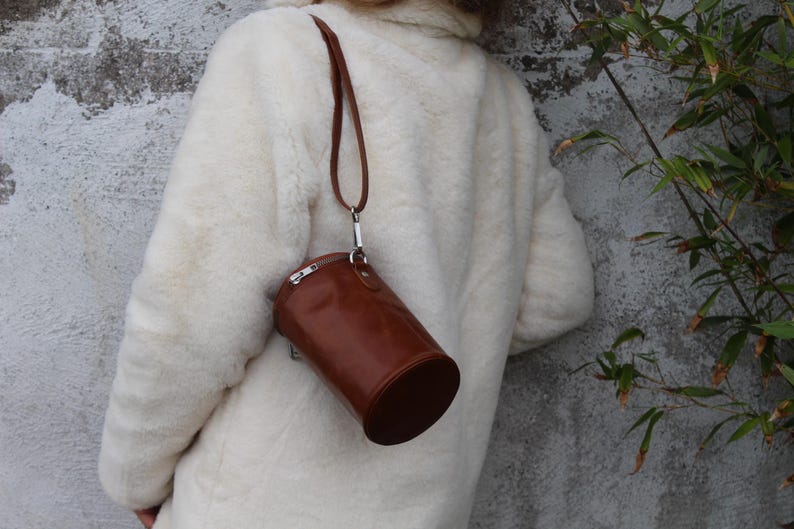 May include: A brown leather cylindrical handbag with a silver zipper and strap is hanging from a shoulder. The bag is hanging against a white fluffy coat. The background is a gray concrete wall.