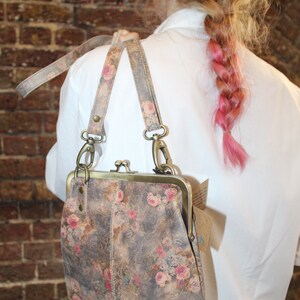 May include: A floral patterned leather purse with a brass clasp and a long strap. The purse is worn over the shoulder of a person with pink hair.