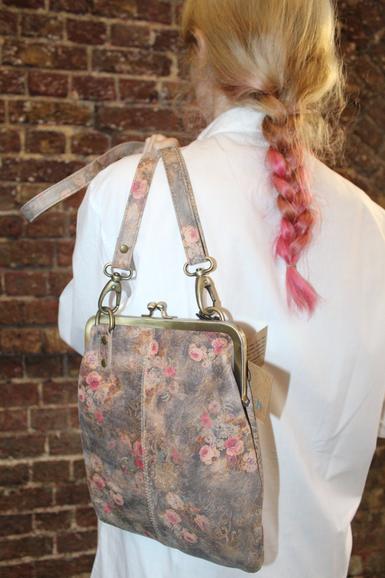 May include: A floral patterned leather purse with a brass clasp and a long strap. The purse is worn over the shoulder of a person with pink hair.