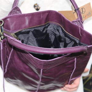 May include: A deep purple leather handbag with a zippered top and a black interior. The bag has a shoulder strap and a small exterior pocket. A tag with the name "Lynch" is attached.