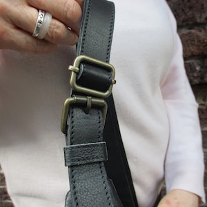May include: Close-up of a black leather shoulder strap with a brass buckle. The strap is adjustable and has a stitched edge.