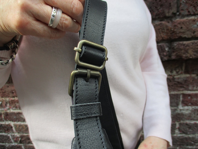 May include: Close-up of a black leather shoulder strap with a brass buckle. The strap is adjustable and has a stitched edge.