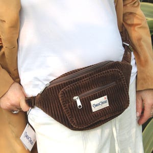May include: A brown corduroy bum bag with two zipped compartments and a silver zip. The bag has a rectangular label with text. The bum bag is worn around the waist.