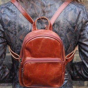 Puede incluir: Una mochila de cuero marrón con un solo bolsillo con cremallera en la parte delantera. La mochila tiene correas ajustables y la lleva puesta una persona.