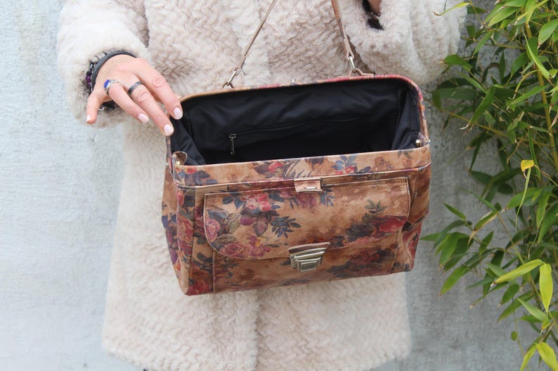 May include: A vintage-style handbag with a floral pattern in shades of brown, red, and pink. The bag has a rectangular shape, a metal clasp, and a black interior. It features a front pocket and a shoulder strap.