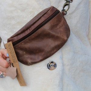 May include: A brown leather fanny pack with a zipper closure and a metal clasp. The bag is worn across the body over a white faux fur coat. A small tag is attached to the bag.