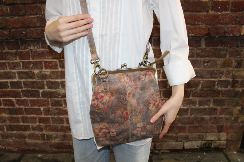 May include: A floral patterned leather crossbody purse with a brass clasp closure and a long adjustable strap.