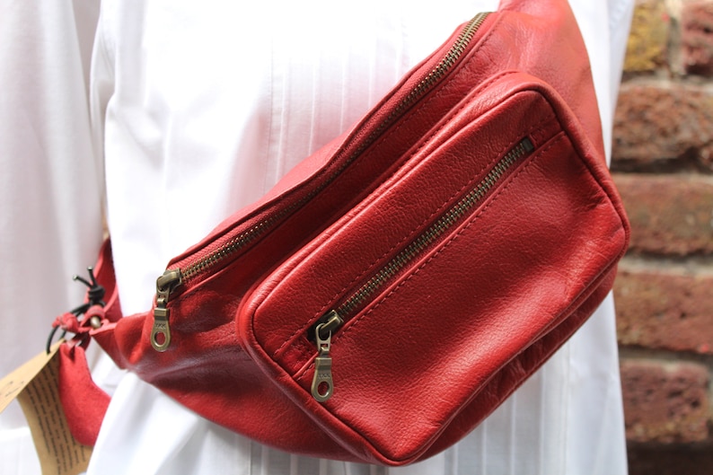 Puede incluir: Una ri&ntilde;onera de cuero roja con dos compartimentos con cremallera. La bolsa se lleva en la cintura.