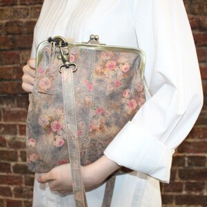 May include: A floral patterned leather purse with a clasp closure and a long strap. The purse is a light brown color with pink and white flowers.