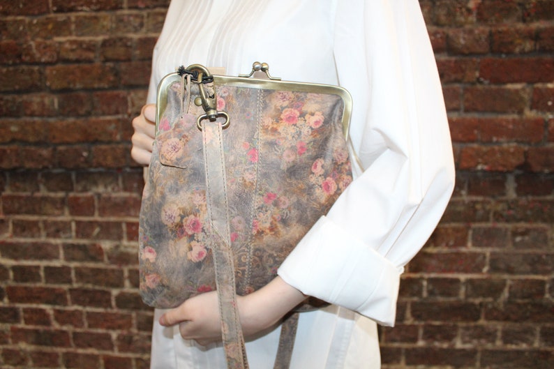 May include: A floral patterned leather purse with a clasp closure and a long strap. The purse is a light brown color with pink and white flowers.