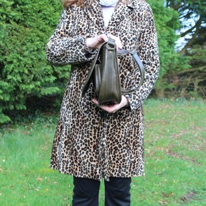 May include: A woman is holding a brown leather satchel bag with a long strap. She is wearing a leopard print coat.
