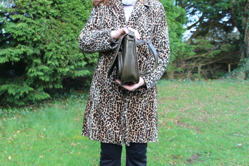 May include: A woman is holding a brown leather satchel bag with a long strap. She is wearing a leopard print coat.