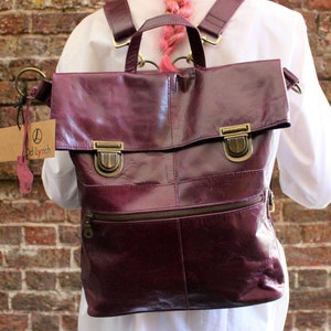 May include: A burgundy leather backpack with a flap closure and brass-coloured hardware. The bag has a front zip pocket and adjustable straps. A tag with the text "D Lynch" hangs from the side. The backpack is worn over a white shirt.