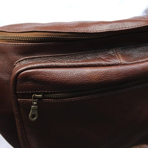 K&ouml;nnte beinhalten: Eine braune Leder-Bauchtasche mit zwei Rei&szlig;verschlussf&auml;chern. Die Tasche hat einen braunen Rei&szlig;verschluss mit einem Metall-Zipper.