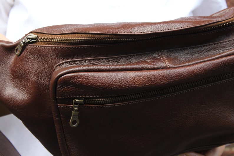 K&ouml;nnte beinhalten: Eine braune Leder-Bauchtasche mit zwei Rei&szlig;verschlussf&auml;chern. Die Tasche hat einen braunen Rei&szlig;verschluss mit einem Metall-Zipper.