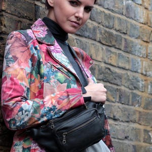 May include: A woman wearing a floral print jacket with a black fanny pack. The jacket has a pink, orange, and green floral pattern. The fanny pack has a zipper closure and a silver buckle.