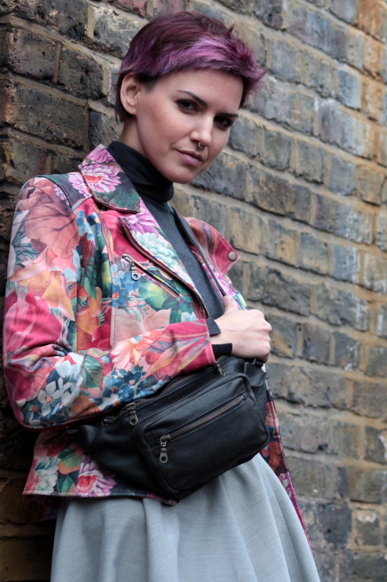May include: A woman wearing a floral print jacket with a black fanny pack. The jacket has a pink, orange, and green floral pattern. The fanny pack has a zipper closure and a silver buckle.