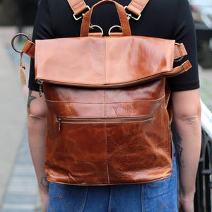 以下が含まれることがあります： フラップ留めと前面にジッパーポケットが付いたブラウンの革製バックパック。バックパックは使い古したような外観で、高品質の革で作られています。