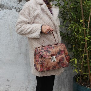 May include: A vintage floral print handbag with a silver clasp. The bag features a brown background with red and pink roses. The handbag is held by a person wearing a cream-coloured coat with a textured, fluffy appearance.
