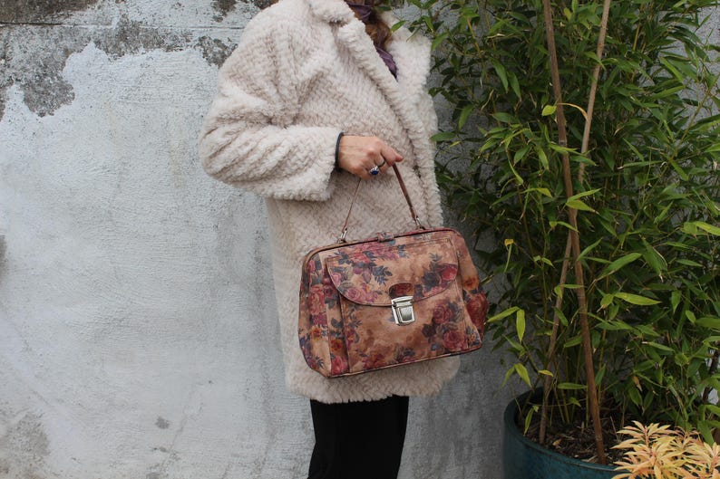 May include: A vintage floral print handbag with a silver clasp. The bag features a brown background with red and pink roses. The handbag is held by a person wearing a cream-coloured coat with a textured, fluffy appearance.