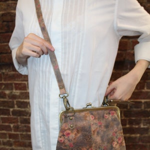 May include: A brown leather crossbody bag with a floral print and a brass clasp. The bag has a long adjustable strap.