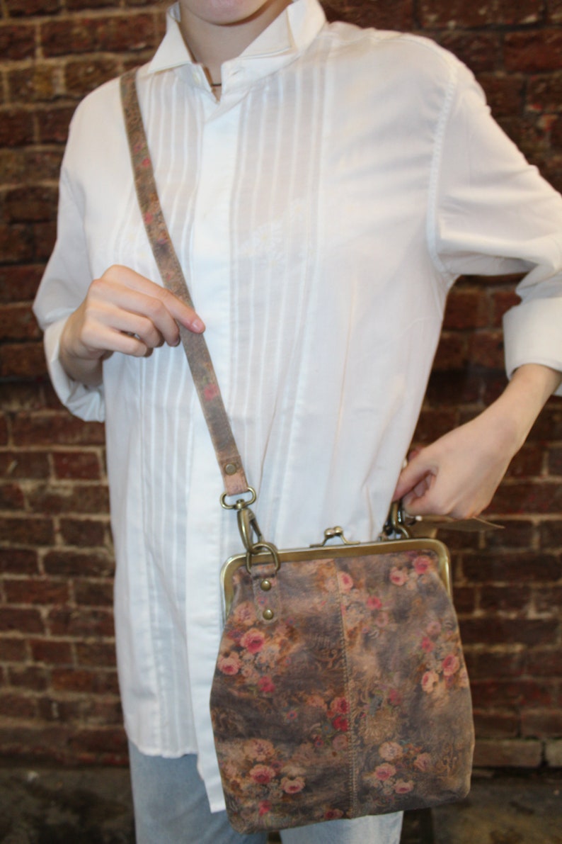 May include: A brown leather crossbody bag with a floral print and a brass clasp. The bag has a long adjustable strap.