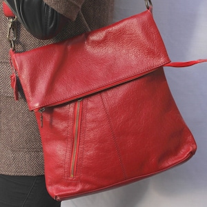 Op de afbeelding: Rode leren crossbodytas met een overslag en een voorvak met ritssluiting. De tas heeft een lange riem en een ritssluiting. Het leer heeft een getextureerde uitstraling.