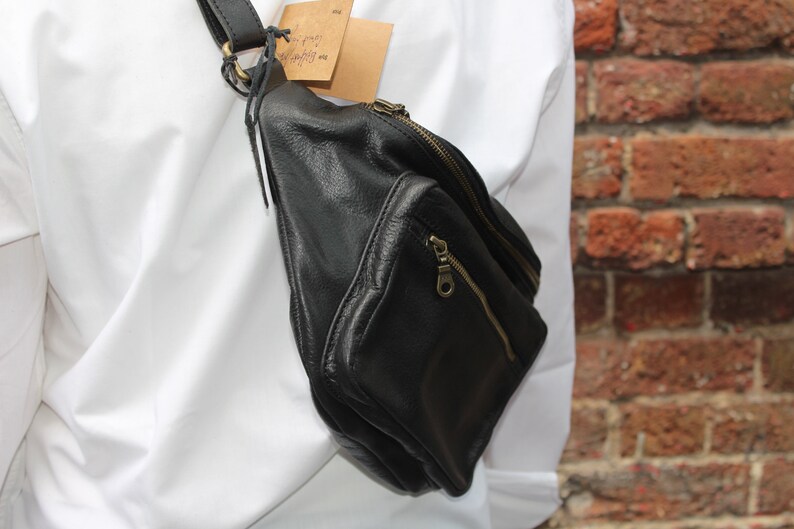 May include: Black leather fanny pack with a zippered main compartment and a smaller front pocket. The bag has a black strap and gold-colored zippers. A small tag is attached to the strap.