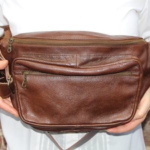 K&ouml;nnte beinhalten: Eine braune Leder-Bauchtasche mit zwei Rei&szlig;verschlussf&auml;chern und einem gro&szlig;en Hauptfach. Die Tasche hat einen verstellbaren braunen Gurt.