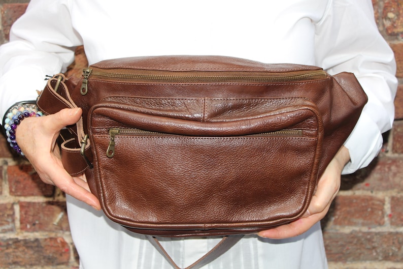 K&ouml;nnte beinhalten: Eine braune Leder-Bauchtasche mit zwei Rei&szlig;verschlussf&auml;chern und einem gro&szlig;en Hauptfach. Die Tasche hat einen verstellbaren braunen Gurt.