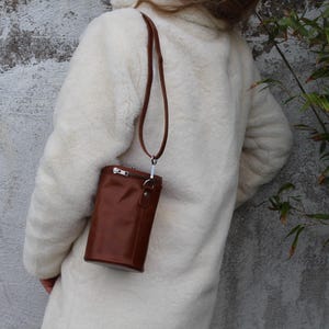 May include: A brown leather crossbody bag with a long strap hangs from the shoulder. The bag has a rectangular shape with a zipper closure and a silver-colored metal clasp. The person is wearing a white coat.