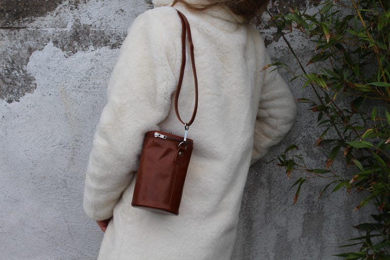 May include: A brown leather crossbody bag with a long strap hangs from the shoulder. The bag has a rectangular shape with a zipper closure and a silver-colored metal clasp. The person is wearing a white coat.